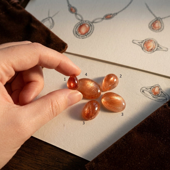 Side view of a hand gently holding a small Oregon Sunstone cabochon with vibrant hues and Schiller effect, jewelry sketches in warm lighting.
