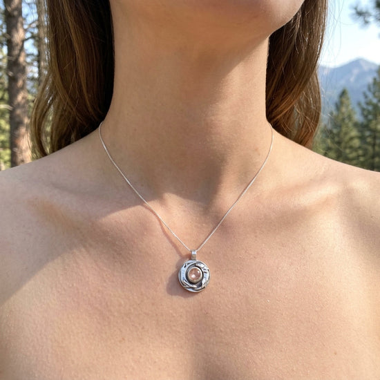 Side view of a model outdoors wearing a tiny silver otter pendant with peach sunstone at her collarbone, in natural daylight with a soft-focus background.