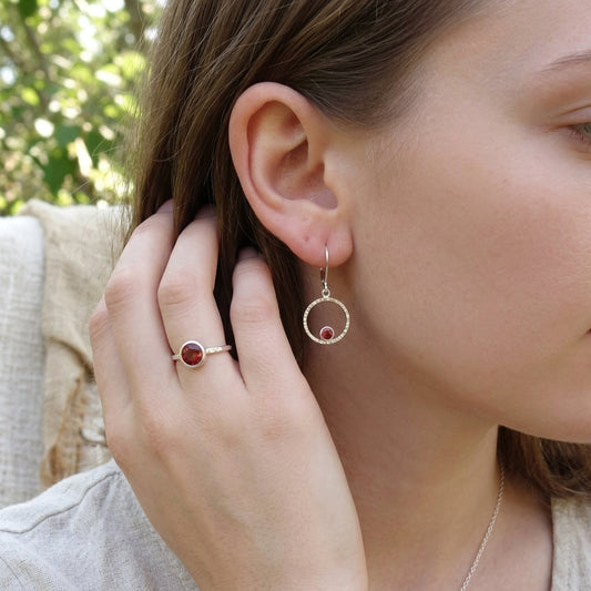 Close-up of a hand wearing small sterling silver Orbit Earrings with Oregon Sunstones, featuring a natural outdoor setting with greenery.