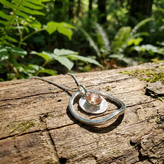 Delicate Sunstar necklace with 5mm Peach Oregon Sunstone pendant on wooden surface with green foliage and sunlight filtering through leaves.