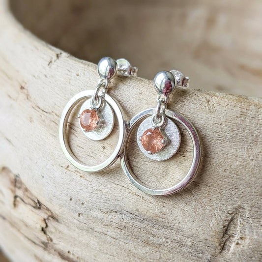 Close-up of Sunstar earrings with sterling silver and peach Oregon sunstone on natural wood background, emphasizing sparkle and textured details.