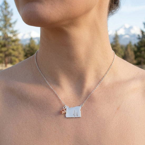 Close-up of small sunstone ring on model's hand, outdoor background with pine trees and snowy mountains, natural soft lighting.
