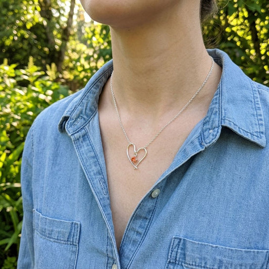 Female model wearing Valentine Heart Necklace with Oregon Sunstone, outdoors with greenery, showcasing delicate jewelry on her neck.