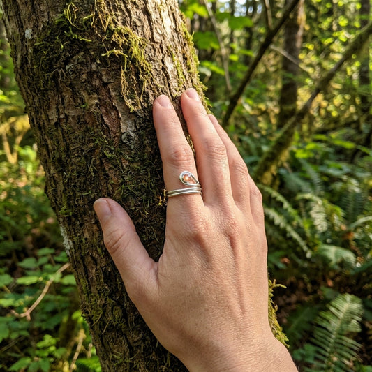 Over-the-shoulder shot of a woman’s hand with Oregon Sunstone ring on a tree trunk, in a forest setting, highlighting craftsmanship.